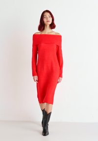Abito rosso a maglia, scollato sulle spalle, con maniche lunghe, silhouette aderente, lunghezza a metà polpaccio e texture a coste attorno al collo. Stivali neri al ginocchio.