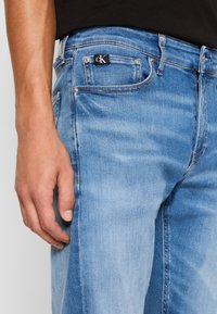 Světle modré džíny z denimu s přiléhavým střihem, viditelnými švy a malou černou etiketou "cK" na zadním kapse.