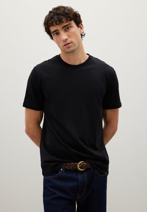 Camiseta de algodón negra con mangas cortas, escote redondo y textura suave, combinada con jeans de mezclilla oscura y un cinturón trenzado marrón.