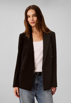 Femme aux longs cheveux bruns portant un blazer noir croisé sur un top blanc et un jean bleu, sur un fond gris uni.