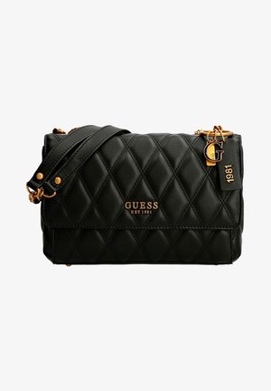 Sac en cuir matelassé noir porté en bandoulière avec une chaîne en or, logo doré "Guess EST 1981" et breloques décoratives dorées en forme de "G" et "1981".