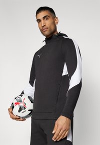 Zwarte pullover hoodie met witte zijkanten, voorzien van een ritskraag en het Puma-logo. Model houdt een zwart-wit voetbal vast.