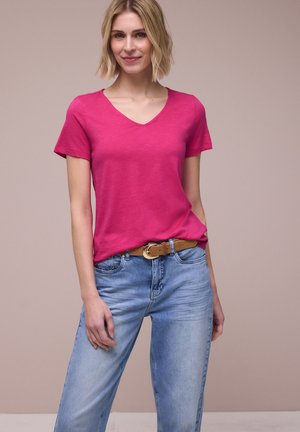 Femme aux cheveux blonds courts portant un t-shirt rose à col en V, rentré dans un jean bleu clair avec une ceinture marron, debout sur un fond uni.