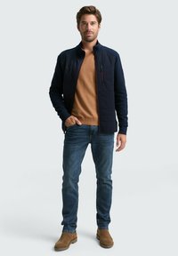 Marineblau gesteppte Jacke mit Stehkragen, kombiniert mit einem hellbraunen Pullover, blauen Jeans und braunen Stiefeletten. Mit Reißverschlusstaschen und einer glatten Textur.