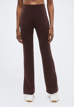 Femme portant un pantalon évasé marron foncé taille haute avec des baskets blanches, debout devant un fond blanc uni.