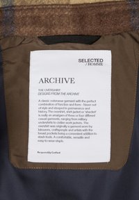 Brun og marineblå overskjorte med en blød tekstur. Har et stofmærke med produktdetaljer, herunder lommer og designbeskrivelse.
