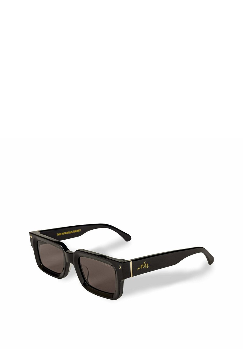 Zwarte rechthoekige zonnebril met donkere lenzen, met gouden accenten op het frame en de armen. Tekst op de arm leest "DE INFAME GURU."