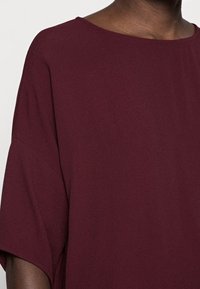 Bordeaux losse-top met een ronde halslijn en korte mouwen. Zachte textuur, subtiele drapering, minimale stikdetails.