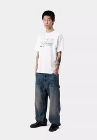 Biała bawełniana koszulka z graficznym nadrukiem z przodu, w połączeniu z szerokimi niebieskimi dżinsami. Model nosi czarne buty o gładkiej teksturze.