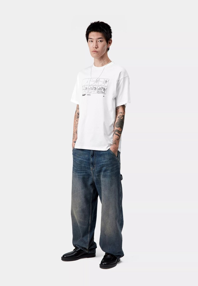 Biała bawełniana koszulka z graficznym nadrukiem z przodu, w połączeniu z szerokimi niebieskimi dżinsami. Model nosi czarne buty o gładkiej teksturze.