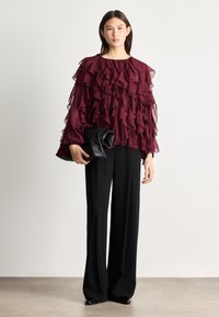 Burgundy ruffled blouse with long sheer sleeves, paired with high-waisted black wide-leg pants. The model holds a black clutch.