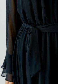Schwarzes, plissiertes Kleid mit fließender Silhouette, das einen gebundenen Taillengürtel und halbtransparente lange Ärmel für einen texturierten Kontrast aufweist.