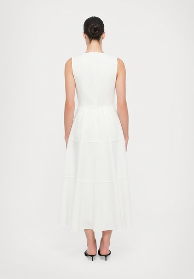 White sleeveless maxi dress with a tiered skirt design, featuring a single back zipper and smooth texture. Black heeled shoes visible.