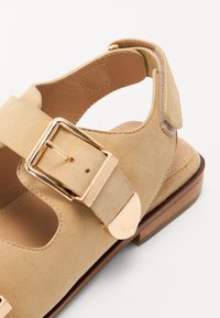 Sandală bej din suede cu brâu pe gleznă, cataramă din aur, design cu decupaje și talpă plată din lemn. Interiorul căptușit este neted.