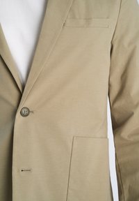 Μπεζ σακάκι με ένα κουμπί, γιακά με σχισμή, τσέπη στο στήθος και μπροστινή τσέπη φορεμένο πάνω από ένα λευκό πουκάμισο.