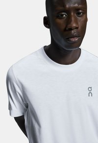 Weißes Baumwoll-T-Shirt mit rundem Ausschnitt, kurzen Ärmeln und einem kleinen grauen Logo auf der Brust. Glatte Textur und lässige Passform.