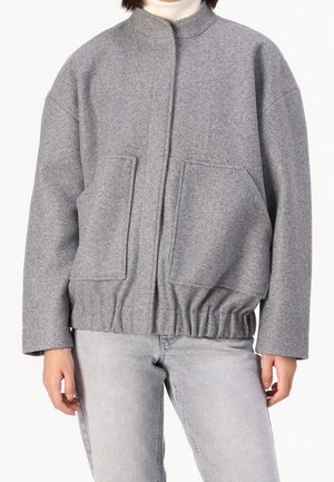 Person trägt eine graue Wolljacke mit großen Fronttaschen und elastischem Bund, kombiniert mit hellgrauen Jeans und einem weißen Rollkragenpullover.