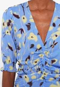 Robe bleue à fleurs jaunes et marron, froncée au centre, encolure en V et manches courtes bouffantes avec poignets élastiques.