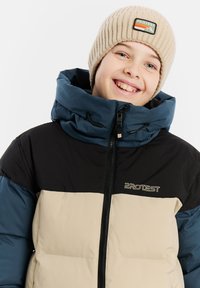 Puffermantel mit schwarzen, beigen und türkisfarbenen Bereichen, hohem Kragen, Frontreißverschluss und Logodetail. Gerippte beigen Strickmütze mit Patch.