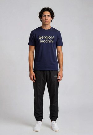 Joven de pie, vistiendo camiseta azul marino con el logo de Sergio Tacchini, pantalones deportivos negros y zapatillas blancas sobre un fondo liso claro.