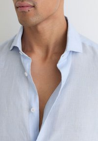 Mężczyzna noszący jasnoniebieską lnianą koszulę z rozpiętym kołnierzykiem, ukazujący szyję, dolną część twarzy i część klatki piersiowej na jednolitym tle.