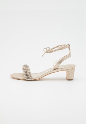 Sandale dorée avec une bride texturée ornée de strass, bout ouvert, et un talon épais de hauteur moyenne ; lien à la cheville pour un ajustement personnalisable.