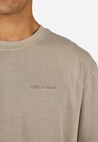 Béžové bavlnené tričko s okrúhlym výstrihom a krátkymi rukávmi. Obsahuje nenápadné vyšívanie loga v zodpovedajúcej farbe na hrudi.