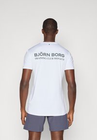 Λευκό αθλητικό μπλουζάκι κοντομάνικο με σκούρο πράσινο κείμενο στην πλάτη: "BJÖRN BORG" και "TRAINING CLUB MONACO". Λείο ύφασμα, εφαρμοστή σχεδίαση.