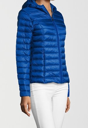 Frau trägt eine blaue gesteppte Kapuzenjacke mit Reißverschluss, kombiniert mit weißen, eng anliegenden Hosen vor einem einfarbigen Hintergrund.