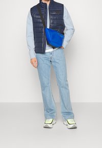 Marinblå vadderad väst, ljusblå sweatshirt och ljusa jeans. En blå axelväska och vita sneakers med gröna detaljer fulländar outfiten.