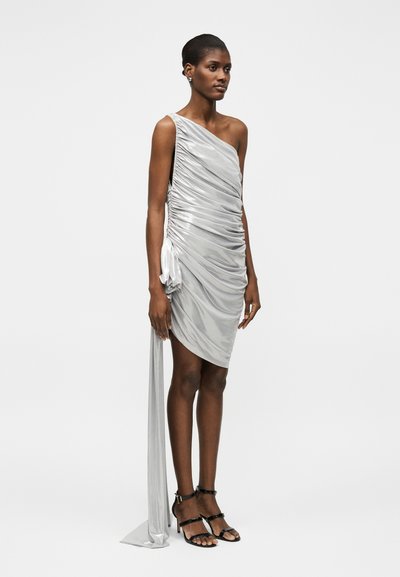 Γυναίκα φοράει αμάνικο, ασημί φόρεμα με έναν ώμο, ραφές και μακριά υφασμάτινη ουρά, σε συνδυασμό με μαύρα πέδιλα με ψηλό τακούνι.