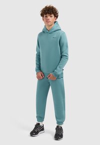 Lichtblauwe hoodie en bijpassende joggingbroek van zachte stof. Heeft een kangoeroezak en logo-opdruk. Gemaakt met zwarte sportschoenen.