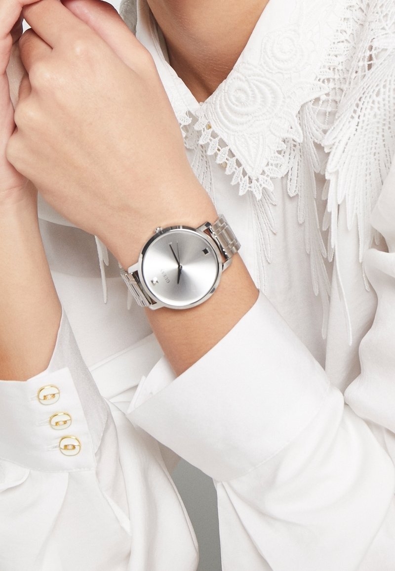 Montre-bracelet avec un bracelet en maille argentée et un cadran argenté minimaliste portée au poignet gauche, associée à une chemise blanche avec col en dentelle et boutons de manchette dorés.