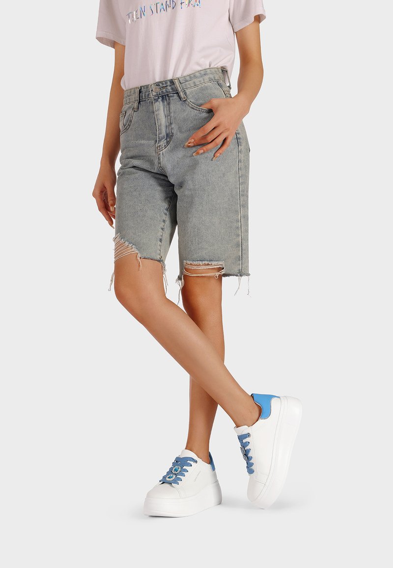 Shorts in denim grigio chiaro con orli sfrangiati, abbinati a sneaker bianche con plateau, caratterizzate da accenti blu e lacci decorativi.