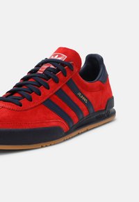 Baskets Adidas en daim rouge avec des bandes noires, des lacets noirs, une semelle en gomme et "JEANS" écrit en or sur le côté.