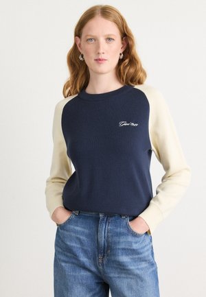 Navy and cream sweatshirt with raglan sleeves and embroidered logo. Soft cotton-blend fabric, worn with high-waisted jeans.