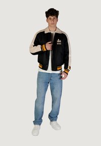 Giacca bomber nera e beige con dettagli arancioni, caratterizzata da un logo sul davanti e polsini a coste, abbinata a jeans azzurri chiari e sneakers bianche.