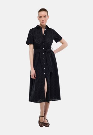 Femme debout, main sur la hanche, portant une robe chemise noire longueur genou avec un motif à œillets et des chaussures Mary Jane tissées foncées.
