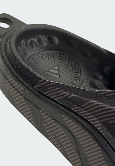 Black slip-on shoe with textured pattern, featuring a cushioned sole, raised dots, and a branded logo in the center.
