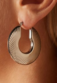 Zilveren oorring met een ribbelstructuur, halfcirkelvorm en een veilige sluiting. Close-upbeeld tegen een huidskleurige achtergrond.