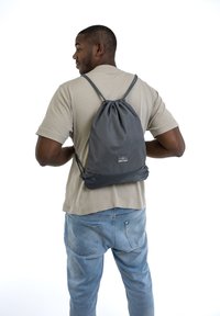 Mochila con cuerda en gris oscuro, con una textura suave y un parche de logo. Se lleva en la espalda, con tirantes ajustables.