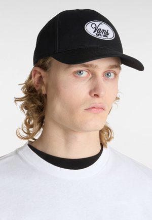 Casquette de baseball noire avec un écusson ovale brodé blanc affichant "Vans" et "EST. 1986." Conçue avec une visière courbée et une texture en tissu.