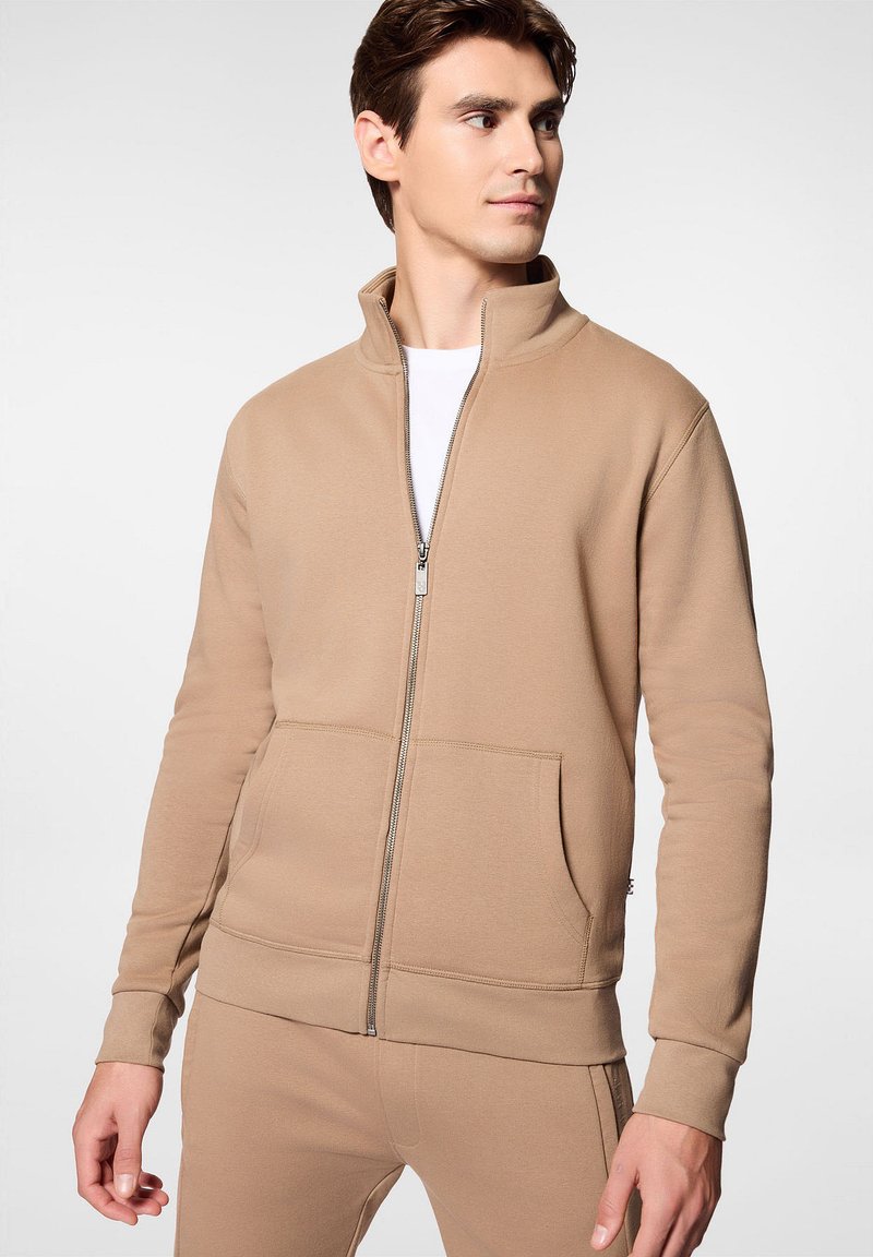 Beżowa bluza z zamkiem na wysokości szyi, z wysokim kołnierzem, dwiema przednimi kieszeniami i ściągaczami na mankietach. Gładki materiał o dopasowanym kroju. W zestawie z pasującymi spodniami.