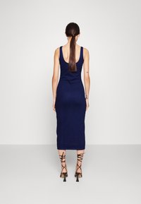 Kvinna med långt mörkt hår i en låg hästsvans, klädd i en ärmlös mörkblå bodycon-klänning och svarta snörade högklackade skor, stående mot en neutral bakgrund.