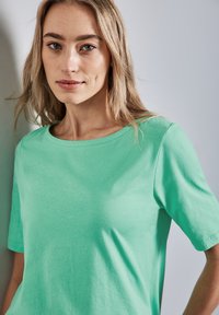 Světle zelené tričko s krátkým rukávem vyrobené z měkkého materiálu, s kulatým výstřihem a volným střihem, které má minimální švy a hladkou texturu.