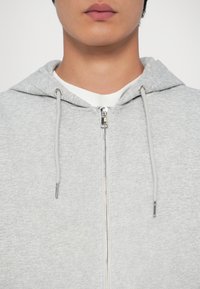 Sudadera con cremallera en color gris claro, con capucha ajustable, cremallera metálica y textura suave. Capa interior blanca visible en el escote.