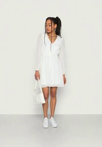 Mladá žena v bielych šatách s výstrihom do V a priehľadnými rukávmi, v bielych teniskách, so skladajúcimi sa náhrdelníkmi a bielou kabelkou, stojaca oproti jednoduchej stene.