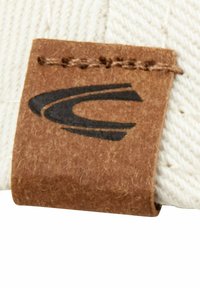 Brun stofetiket med sort logo, syet fast på en lysfarvet tekstiloverflade, som har et struktureret, vævet design.