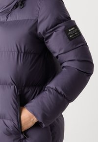 Paarse pufferjack met een geborduurd patroon, een ritszak en een stoffen label op de mouw die de materiaal samenstelling vermeldt.