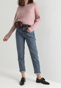 Rosa gestrickter Pullover, hoch taillierte blaue Jeans aus Denim, schwarze Loafers mit Schnalle und eine schwarze, gemusterte Bauchtasche, die in der Taille getragen wird.